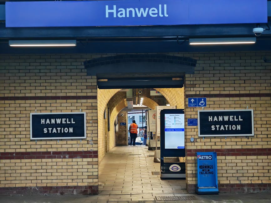 The entrance to Hanwell Station is framed by a brick archway with cream and brown bricks, leading into a walkway with tiled flooring. To the left and right of the arch, there are two black and white s